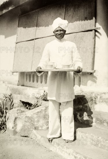 An Indian house servant. An Indian house servant poses on the steps of a colonial bungalow holding a tray set with a china teapot and cups. Chikmagalur, Mysore State (Chikkamagaluru, Karnataka), India, circa 1936., Karnataka, India, Southern Asia, Asia.