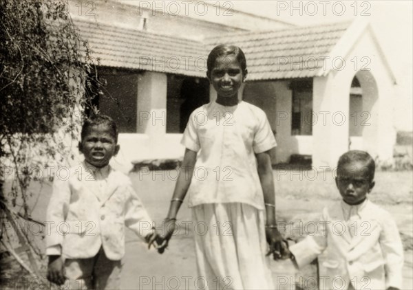 Indian siblings in Western-style dress. Portrait of three Indian siblings in Western-style dress holding hands on the front lawn of a colonial bungalow. Originally captioned as 'T. Devadatta's children', they are the children of an evangelist living in a Methodist mission compound. Gubbi, Mysore State (Karnataka), India, 1933., Karnataka, India, Southern Asia, Asia.