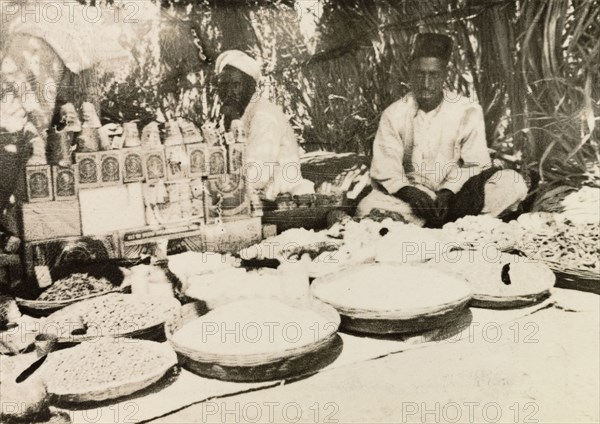 Food stall at Indian market. Two street traders sit in the shade of their covered market stall, which sells a variety of food displayed in shallow baskets placed on the ground. Mysore State (Karnataka), India, 1933., Karnataka, India, Southern Asia, Asia.