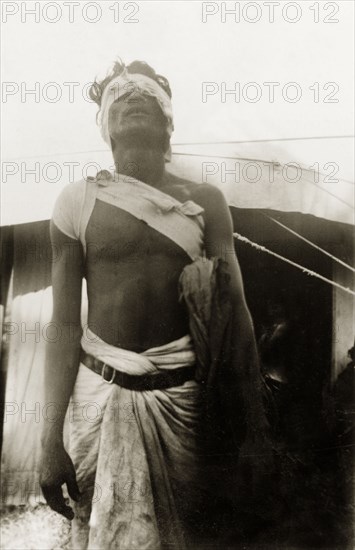 Patient at a missionary medical camp. A male patient at a Methodist mission medical camp emerges from a tent with bandages wrapped round his head and shoulder. Mysore State (Karnataka), India, 1934., Karnataka, India, Southern Asia, Asia.