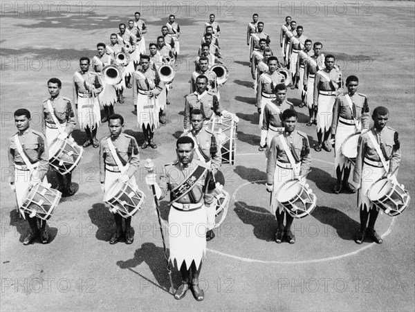 Royal Fijian Military Band. Fronted by a Drum Major holding a staff, the 32-piece Royal Fijian Military Band stands in formation, ready to march. The band's instruments include drums, trombones, tubas and trumpets. Fiji, 1965. Fiji, Pacific Ocean, Oceania.
