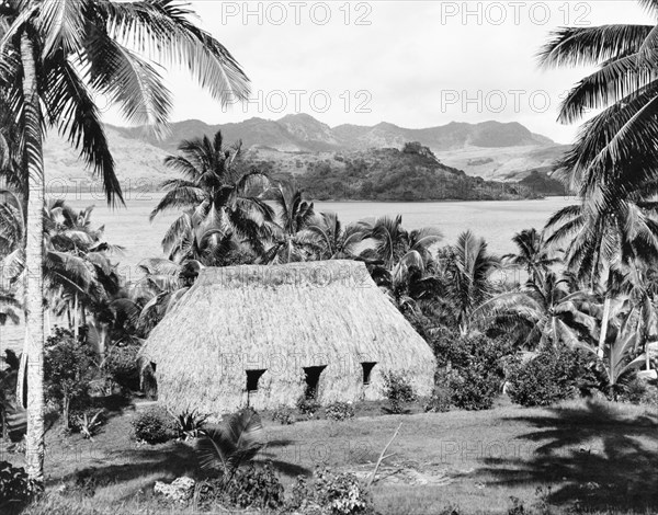 A traditional Fijian 'bure'. A traditional Fijian 'bure' (dwelling) sits amongst palm trees at the bottom of a slope, overlooking a coastal bay. Bures such as this usually consist of a wooden frame, which is completed covered with woven leaves and thatch. Fiji, 1965. Fiji, Pacific Ocean, Oceania.