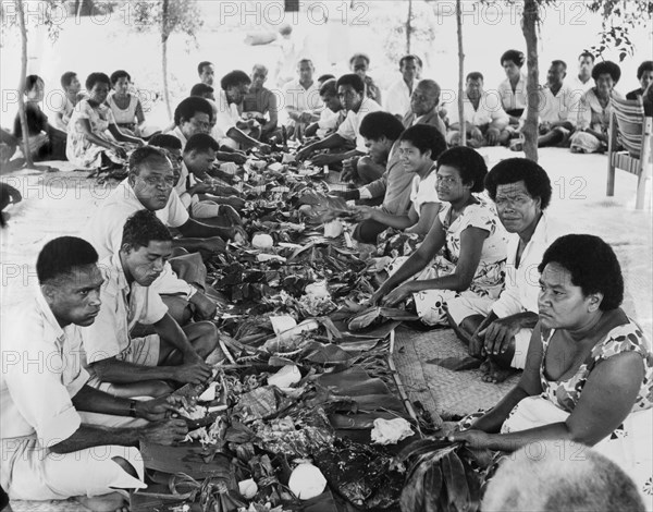 A Fijian 'magiti'. Two lines of diners sit cross-legged on the ground opposite each other during a 'magiti' (Fijian feast). Between them are plaited coconut leaf platters on which has been placed food wrapped in banana leaves. An original caption comments: "The food, most of which has been cooked in 'lovos' (ground ovens), varies from prawns, fish, crabs, pork and beef, to vegetable dishes and Fijian puddings". Fiji, 1965. Fiji, Pacific Ocean, Oceania.
