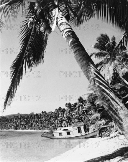 A beached boat, Fiji. A small motor boat lies stricken on a sandy beach, after having its ?bottom ripped out on a hidden reef". Fiji, 1965. Fiji, Pacific Ocean, Oceania.