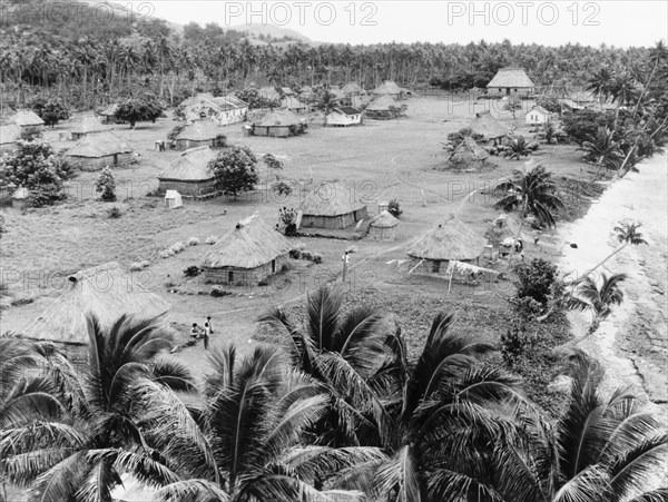 A 'bure' village in Fiji. A village comprising a number of traditional Fijian 'bures' (dwellings) sits in a palm forest clearing beside a beach. Fiji, 1965., Fiji, Pacific Ocean, Oceania.