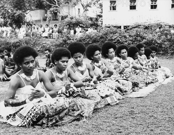 Women perform a 'vakamalolo'. Ten young Fijian women sit cross-legged in a line and clasp their hands together as they perform a 'vakamalolo' or sitting dance outdoors. All wear ceremonial costume including patterned 'sulus' (wraparound skirts) and typical Fijian hairstyles. Fiji, 1965. Fiji, Pacific Ocean, Oceania.