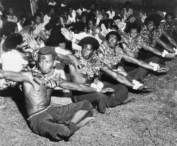 Men perform a 'vakamalolo'. A group of young Fijian men sit cross-legged in a line and stretch out their arms as they perform a 'vakamalolo' or sitting dance. Naked from the waist up, they wear ceremonial costume including face paint and leis made from leaves. Fiji, 1965. Fiji, Pacific Ocean, Oceania.