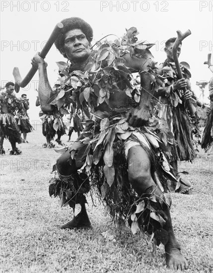 Men perform the 'meke i wau'. A group of men perform the 'meke i wau', a male dance from Fiji incorporating clubs. They dance wearing ceremonial costumes made from grass and leaves. An original caption comments: "The movements are always vigorous, for they portray phases of tribal battles". Fiji, 1965. Fiji, Pacific Ocean, Oceania.