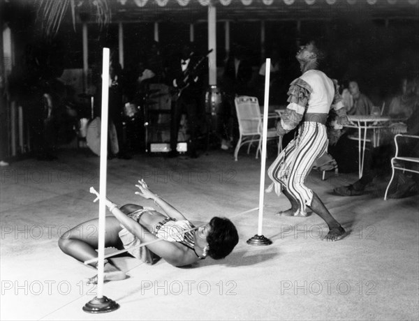 Limbo dancing at the Anchorage Hotel. A female limbo dancer passes beneath a horizontal pole suspended only 20 centimetres from the ground whilst her male partner dances around her, accompanied by a musical band. The couple were cabaret entertainers at the Anchorage Hotel and a related caption comments: ?Ruth was once a schoolmistress. Now she earns a good salary as a limbo dancer?. St Johns, Antigua, 1965. St John's, St John (Antigua and Barbuda), Antigua and Barbuda, Caribbean, North America .