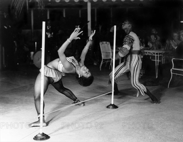 Limbo dancing at the Anchorage Hotel. A female limbo dancer bends over a limbo pole backwards whilst her male partner dances around her, accompanied by a musical band. The couple were cabaret entertainers at the Anchorage Hotel and a related caption comments: ?Ruth was once a schoolmistress. Now she earns a good salary as a limbo dancer?. St Johns, Antigua, 1965. St John's, St John (Antigua and Barbuda), Antigua and Barbuda, Caribbean, North America .