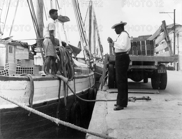 Policing the Careenage. A harbour police officer talks to a man unloading bricks from a sail boat onto a truck as he patrols the Careenage. An original caption comments: "Harbour policemen in the (sailor's) uniform of... Nelson's day are always a target for photographers". Bridgetown, Barbados, 1965. Bridgetown, St Michael, Barbados, Caribbean, North America .