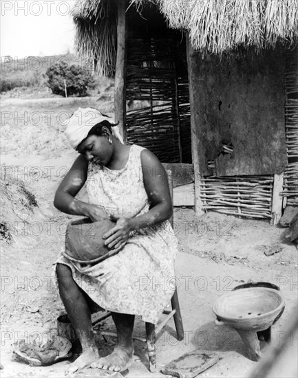 A potter in Monserrat. A woman fashions a clay pot by hand, resting it on her lap as she sits outside a thatched hut. Montserrat, 1965. Montserrat, Caribbean, North America .