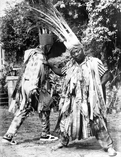 Carnival dancers on Nevis. Two men perform a carnival dance outdoors. Both wear crowns adorned with peacock feathers and costumes made from ribbons and scarves. Nevis, 1965., St Kitts and Nevis, Caribbean, North America .