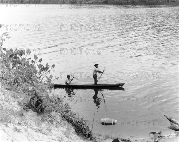 Traditional fishing in British Guiana. Two fishermen prepare their bows and arrows as they pilot a canoe along a wide river. British Guiana (Guyana), 1965., Guyana, South America, South America .