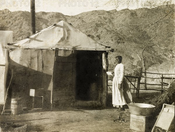 Living quarters at Red Cloud Mine. An African woman wearing an apron stands at the doorway of a canvas tent: part of the living quarters at Red Cloud gold mine in the Chuckwalla Mountains. A large tin bowl and a washboard are placed in the foreground. Southern California, United States of America, March 1902., California, United States of America, North America, North America .