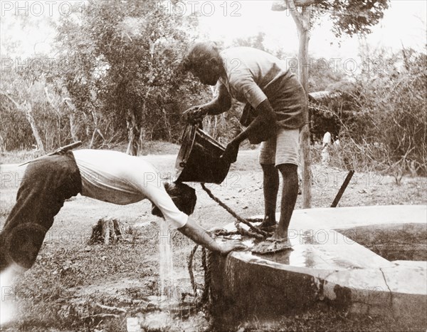Showering at a well, Ceylon. A Ceylonian man pours a bucket of water from a well over a European man's head, providing him with a refreshing shower. Colombo, Ceylon (Sri Lanka), November 1902. Colombo, West (Sri Lanka), Sri Lanka, Southern Asia, Asia.