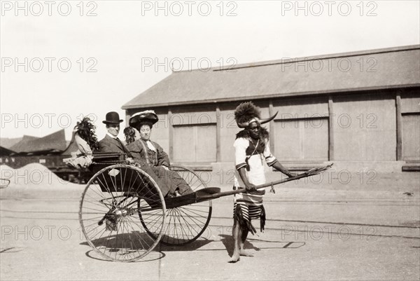 Zulu rickshaw driver, Durban. A Zulu rickshaw driver takes a European couple for a ride. He is dressed in a traditional costume with a flamboyantly horned and feathered headdress. Durban, South Africa, 1904. Durban, KwaZulu Natal, South Africa, Southern Africa, Africa.