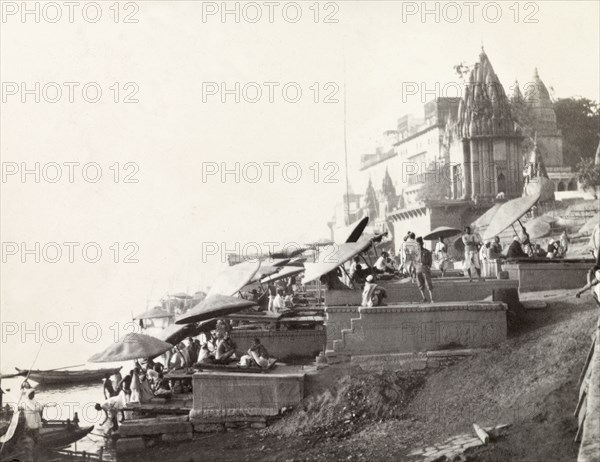 Ghats at Benares . Religious pilgrims sit beneath large umbrellas at a ghat (stepped wharf) on the banks of the River Ganges at Benares, one of Hinduism's holiest sites. The ornate roof of a large Hindu temple can be seen in the background. Benares, United Provinces (Varanasi, Uttar Pradesh), India, December 1902. Varanasi, Uttar Pradesh, India, Southern Asia, Asia.
