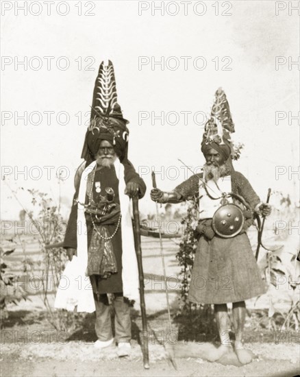 Akalis at Coronation Durbar, 1903. Full-length portrait of two Akalis (Nihang), prestigious Sikh warriors, who wear traditional, tall turbans coiled with steel, in which to store weapons such as knives, swords, and kirpans (Sikh ceremonial daggers). These Akalis had travelled from the Harimandir Sahib (Golden Temple) in Amritsar, Punjab, to Delhi to attend Edward VII's Coronation Durbar. Delhi, India, circa 1 January 1903. Delhi, Delhi, India, Southern Asia, Asia.