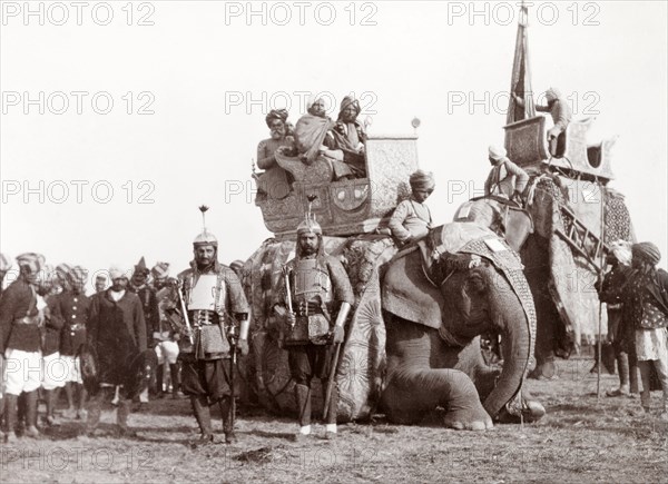 Bhopal State procession at Coronation Durbar, 1903. Two armoured soldiers of Bhopal State stand beside a kneeling caparisoned elephant, on whose back three Indian dignitaries are sitting in an ornate howdah. A second elephant approaches the group, ready to pick up its passengers and join a procession at Edward VII's Coronation Durbar. Delhi, India, circa 1 January 1903. Delhi, Delhi, India, Southern Asia, Asia.