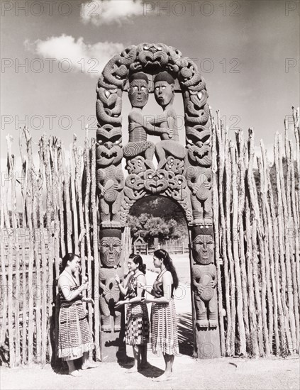 Women beneath a Maori arch. Three young Maori woman in traditional dress chat beneath an ornate Maori arch that has been elaborately carved with wooden figures and swirling patterns. Rotorua, New Zealand, 1966. Rotorua, Bay of Plenty, New Zealand, New Zealand, Oceania.