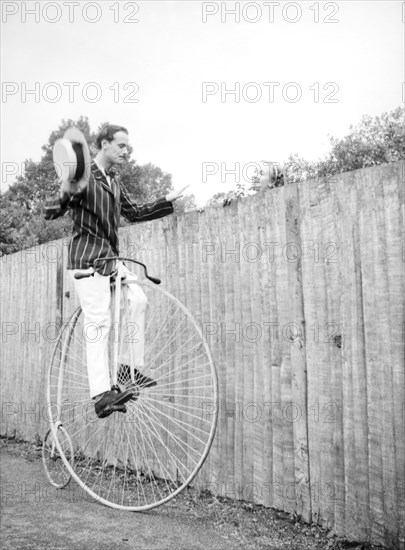 Look, no hands. A woman peers over the top of a tall fence to watch a man in a striped jacket riding a penny-farthing bicycle with no hands. Related information suggests the pair were involved in making a short film with friends. Eastbourne, England, circa 1939. Eastbourne, Sussex, England (United Kingdom), Western Europe, Europe .