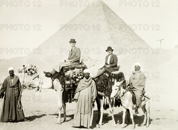 Riding camels at the Great Pyramid. William Ryder McGeagh, District Commissioner in Jerusalem, and his wife ride a pair of caparisoned camels in front of the Great Pyramid. They are guided by three Egyptian men in long robes: one of whom rides a donkey. Giza, Egypt, January 1939. Giza, Giza, Egypt, Northern Africa, Africa.