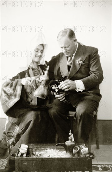 Portrait of Mr and Mrs Keith-Roach . Edward Keith-Roach, District Governor of Jerusalem, holds out a small ceramic cup as his wife pours him a drink of coffee from an ornate metal jug. A second jug sits in a trunk of hot ashes on the floor. Related photographs show that the couple had recently been married in a traditional Christian ceremony, but Keith-Roach's wife appears here wearing a traditional Palestinian 'thob'. Jerusalem, British Mandate of Palestine (Israel), circa 1939., Bedfordshire, England (United Kingdom), Western Europe, Europe .