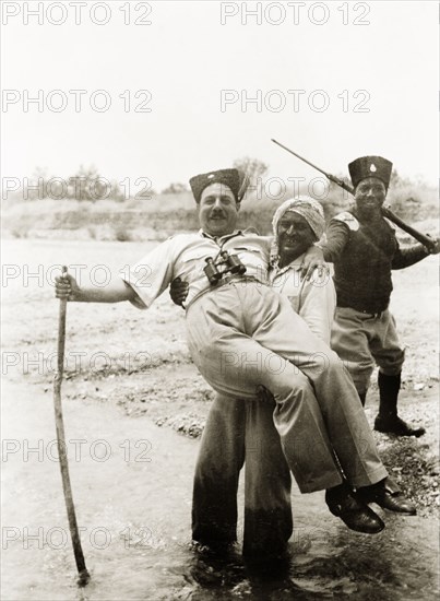 A hike in the Palestinian countryside. An Arab guide stops to smile for the camera as he carries a European man across a stream during a country hike. Behind them is a uniformed guard with a rifle, who appears to be of African descent. British Mandate of Palestine (Israel), 1940. Israel, Middle East, Asia.