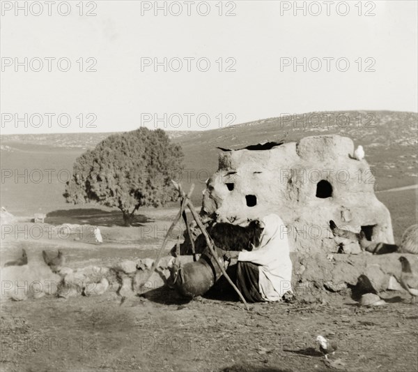 Domestic duties in Palestine. A veiled Arab woman performs some sort of domestic task using a sack suspended from a tripod. Behind her is a large, clay structure, possibly a dovecot or chicken coup. British Mandate of Palestine (Israel), circa 1940. Israel, Middle East, Asia.