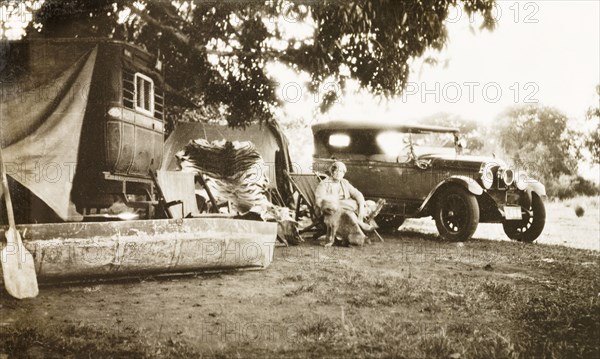Relaxing at a hunting camp, India. A European woman relaxes in a deckchair with her pet dog at a hunting camp in the country. A tiger skin is stretched out behind her and she is surrounded by vehicles including a motor car and a canoe. Mandagadde, Mysore State (Karnataka), India, circa 1935., Karnataka, India, Southern Asia, Asia.
