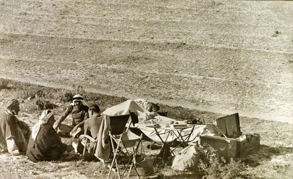 A travelling party in Al-Masfara. A party of travellers stops for lunch during a journey through Al-Masfara, a mountainous region to the west of the Dead Sea. Three Arab men sit on the ground as they talk to a British man in a deckchair. British Mandate of Palestine (West Bank, Middle East), 2-9 April 1941., West Bank, West Bank (Palestine), Middle East, Asia.