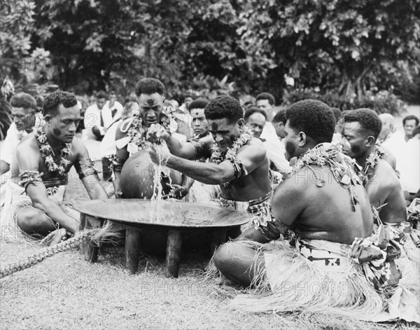 Performing a 'sevusevu' ceremony. Five Fijian men sit in a circle around a wooden 'tanoa' as they participate in a traditional 'sevusevu' welcoming ceremony. Naked from the waist up, the men wear face paint, grass skirts and leis made from leaves. The central figure strains a mixture of water and powdered kava root (Piper methysticum) through a cloth to remove any grit, before distributing the infusion. Fiji, 1965. Fiji, Pacific Ocean, Oceania.