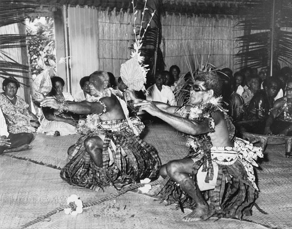Dancing at a 'sevusevu' ceremony. Two Fijian men perform a dance with fans at a traditional 'sevusevu' welcoming ceremony. They wear ceremonial costume including dramatic face paint, skirts made from strips of fabric and leis made from leaves. Behind them is a traditional wooden 'tanoa' (bowl), which is used during the ceremony to mix an infusion of water and powdered kava root (Piper methysticum). Fiji, 1965. Fiji, Pacific Ocean, Oceania.