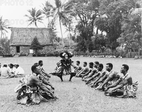 Preparations for a 'sevusevu' ceremony. A group of Fijian men perform an elaborate dance before a traditional 'sevusevu' welcoming ceremony. Most sit cross-legged on the ground, facing each other in two rows, whilst another man dances in the centre, his legs bent and arms outstretched. All wear ceremonial costume including skirts made from palm leaves or strips of fabric. Fiji, 1965. Fiji, Pacific Ocean, Oceania.
