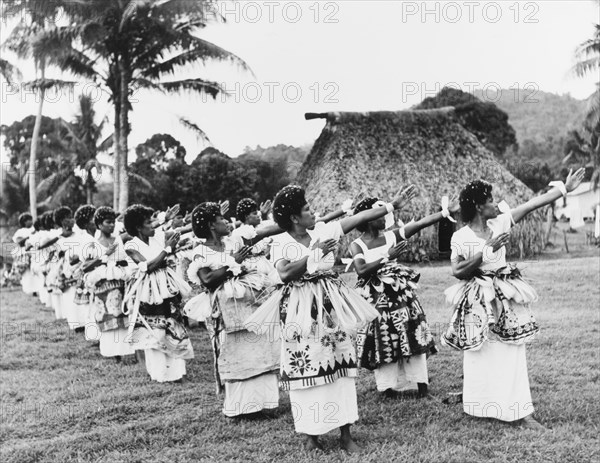 Fijian dancers at Nasilai. A large group of Fijian women perform a traditional dance outdoors. Dressed in ceremonial costume including tiered 'masi' (bark cloth) skirts, they dance in formation, turning their heads and stretching their arms out to one side. Nasilai, Viti Levu, Fiji, 1965. Nasilai, Viti Levu, Fiji, Pacific Ocean, Oceania.