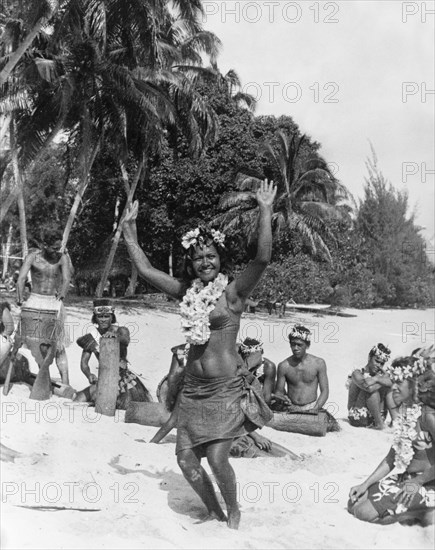 Performers on Moorea Island. A woman holds her hands in the air as she dances on a beach, dressed in a 'pareu' (sarong) and flowered lei. Behind her, several other performers sit in the sand, including four men with traditional Tahitian drums. Moorea Island, Windward Islands, French Polynesia, 1965., Windward Islands (including Tahiti), French Polynesia, Pacific Ocean, Oceania.