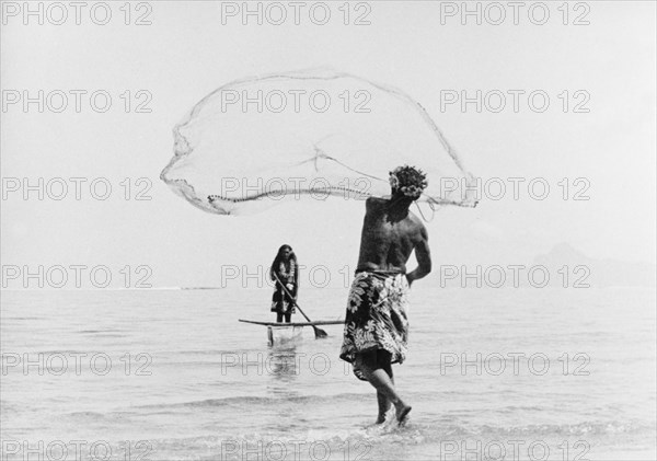 Fishing with a cast net. A Tahitian man wearing a printed 'pareu' (sarong) hurls a cast net into shallow waters off the coast. Opposite him, a woman wearing a flowered lei pilots a canoe equipped with an outrigger. Tahiti, French Polynesia, 1965., Windward Islands (including Tahiti), French Polynesia, Pacific Ocean, Oceania.