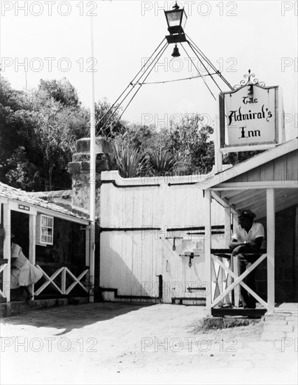 Bolted gate at the Admiral's Inn. A uniformed man leans on a handrail inside a bolted gate. Above him, a sign suggests this may be part of the Admiral's Inn, a historic public house located within Nelson's Dockyard. To the left is a single storey building identified as a guard house. English Harbour, Antigua, 1965. English Harbour, St Paul (Antigua and Barbuda), Antigua and Barbuda, Caribbean, North America .