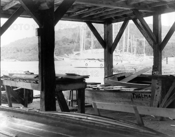 A boat-building workshop at English Harbour. Wooden beams support the roof of a boat-building workshop on the waterfront at English Harbour. In the distance are moored a number of sleek ketches (twin-masted sail boats). English Harbour, Antigua, 1965. English Harbour, St Paul (Antigua and Barbuda), Antigua and Barbuda, Caribbean, North America .
