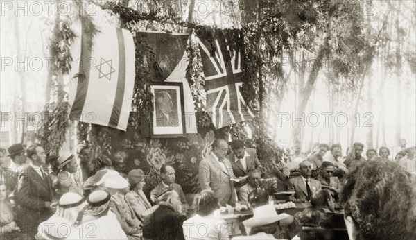 Opening of the Benes Garden in Gan Yavne. A man identified as 'Novak' addresses a seated audience at the opening of the Benes Garden in commemoration of deposed Czech President, Edvard Benes. Behind him hangs a framed portrait of Benes, surrounded by a Jewish flag and a union jack. Gan Yavne, British Mandate of Palestine (Central Israel), 4 June 1944., Central (Israel), Israel, Middle East, Asia.