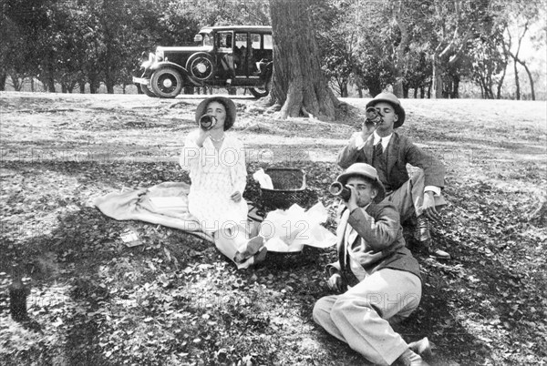 Picnic en route to Bareilly. Two members of the Police Training School and a female friend have a picnic lunch on the road to Bareilly. They sit on the grass, drinking from glass bottles, in front of a motorcar parked in the distance. Near Bareilly, United Provinces (Uttar Pradesh), India, February 1934. Bareilly, Uttar Pradesh, India, Southern Asia, Asia.