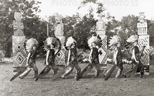 Chorus line act at a garden party. Members of the cast of 'Flying Fragments' performed by the Moradabad Amateur Dramatic Society at the Railway Institute. Several women perform a chorus line act at a garden party, dressed in traditional Native American costume. They strike a pose in front of four artificial totem poles, each holding a large oar with both hands as part of the Red Indian Canoe Dance. Moradabad, United Provinces (Uttar Pradesh), India, 12 February 1934. Moradabad, Uttar Pradesh, India, Southern Asia, Asia.