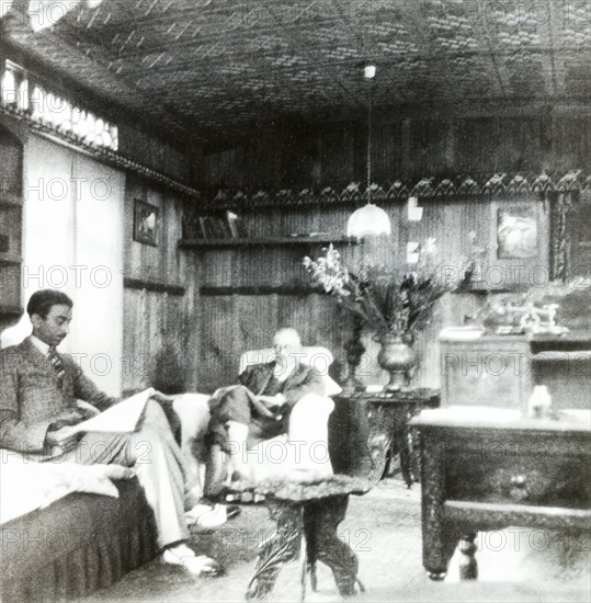 Reading the papers inside a shikara. Two men sit in armchairs, reading newspapers in the panelled sitting room of the houseboat 'Pigeon' at Chenar Bagh. Seated in background is Hugh Inglis, Principal of the Police Training School, Moradabad. Srinagar, Jammu and Kashmir, India, May 1934. Srinagar, Jammu and Kashmir, India, Southern Asia, Asia.