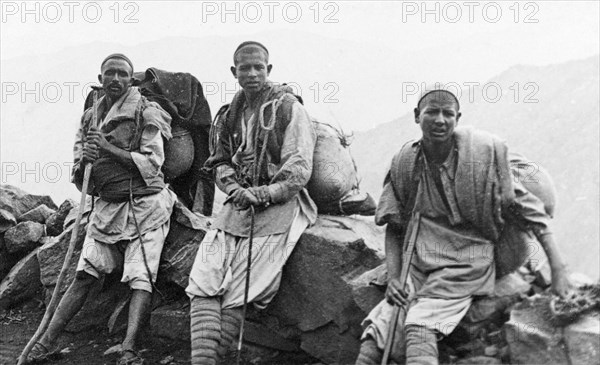 Kashmiri travellers at rest. Three of George Boon's Kashmiri porters rest their heavy loads against a rocky wall on a mountain pass on the way to Shisham Nag. Each carries a blanket and wears several layers of clothing including a kufi (cap) and protective leg puttees (bound strips of cloth). Srinagar, Jammu and Kashmir, India, 1934., Jammu and Kashmir, India, Southern Asia, Asia.