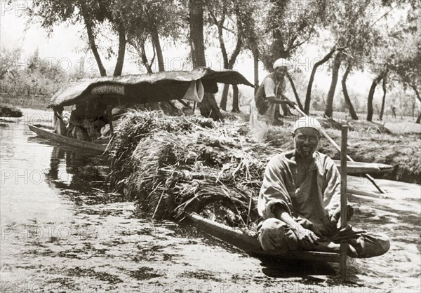 A canal dredger in Srinagar. A canal dredger sits cross-legged on the bow of a long canoe, which is piled high with cut reeds. Srinagar, Jammu and Kashmir, India, 1934. Srinagar, Jammu and Kashmir, India, Southern Asia, Asia.