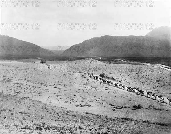 Camel convoy at the entrance to the Khyber Pass. A camel convoy winds its way through the desert towards the entrance of the Khyber Pass. An original caption points out another "road for motor transport" (top right) and a small cemetery located on a hill in the distance. North West Frontier Province, India (Federally Administered Tribal Areas, Pakistan), circa 1919., Federally Administered Tribal Areas, Pakistan, Southern Asia, Asia.