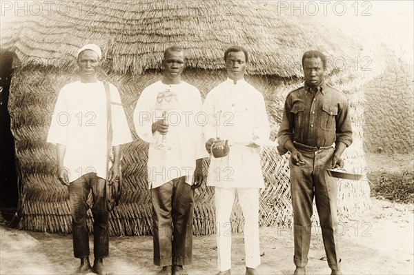 Four Nigerian domestic servants. Portrait of four Nigerian servants standing outside a traditional style, woven hut. They pose for the camera holding various household items including a lamp, frying pan, teapot and saucers. Nigeria, circa 1915. Nigeria, Western Africa, Africa.