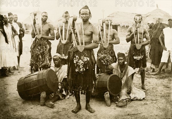 Nigerian musicians and hoe dancers. Five Nigerian hoe dancers, dressed in grass skirts with bells and bangles, pose for a group portrait holding their hoes. Two musicians sit on the floor amongst the group, playing the 'ganga' (drums) with curved sticks. Nigeria, circa 1915. Nigeria, Western Africa, Africa.