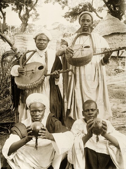 Nigerian musicians with their instruments. Four Nigerian musicians pose for a group portrait with their instruments. Two of the men stand, carrying 'gojes' (one or two-string fiddles) with curved bows, whilst the other two squat on the ground, holding round percussion instruments. Borno State, Nigeria, circa 1915., Borno, Nigeria, Western Africa, Africa.